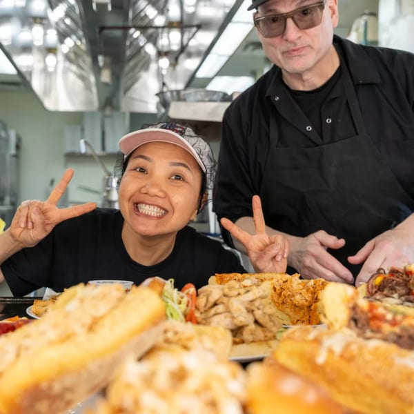 Two chefs, one smiling and making a peace sign, behind a counter laden with food.