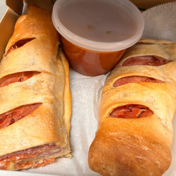 Two baked strombolis with visible meat and cheese filling, alongside a container of red sauce.