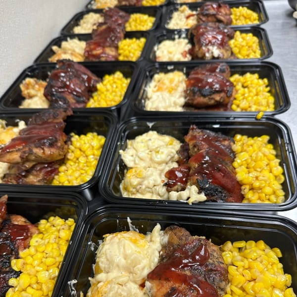Rows of Curly's Comfort Foods BBQ chicken, macaroni and cheese, and corn in black to-go containers.